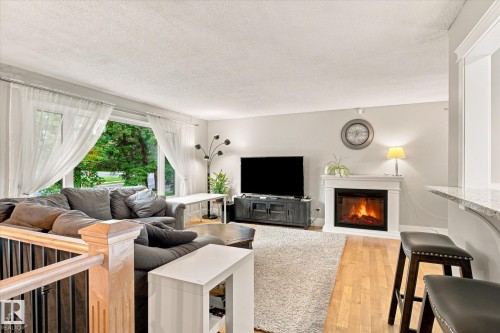 Hardwood-style flooring throughout, featuring a white mantle fireplace with hearth, an integrated breakfast bar with a stone-finish countertop, a large picture window, and a wood-finish staircase with black spindles - 3 Akins Drive, St. Albert, AB 