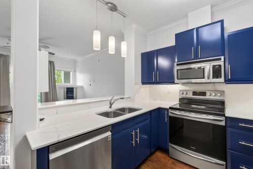 Kitchen featuring blue cabinetry, white countertops, stainless steel appliances, and wood-finish flooring - #210 10121 80 Av Nw, Edmonton, AB 