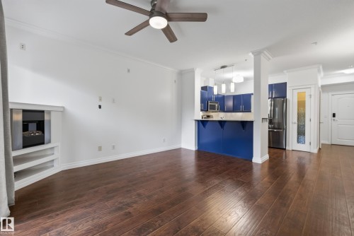 Dark wood-finish flooring with a ceiling fan, a built-in fireplace with shelving, and a kitchen featuring blue cabinetry and stainless steel appliances - #210 10121 80 Av Nw, Edmonton, AB 