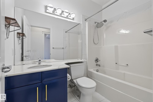 Vanity with a white countertop and integrated sink, blue cabinetry with gold-toned pulls, full-width mirror, and a white bathtub with an integrated shower - #210 10121 80 Av Nw, Edmonton, AB 