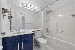 Vanity with a white countertop and integrated sink, blue cabinetry with gold-toned pulls, full-width mirror, and a white bathtub with an integrated shower - 
