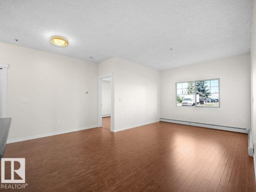 Expansive living area featuring wood-finish flooring, neutral wall tones, a large window, and white trim throughout - #116 16807 100 Av Nw, Edmonton, AB 