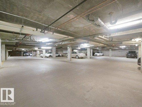 Underground parking facility with a concrete ceiling and flooring, featuring ample lighting, exposed piping, and structural support columns - #116 16807 100 Av Nw, Edmonton, AB 
