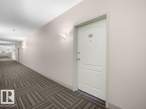 Hallway with patterned carpeting, white wall sconces, and a paneled entry door - #116 16807 100 Av Nw, Edmonton, AB 