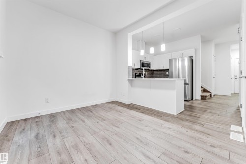 Open-concept kitchen with white cabinetry and stainless steel appliances, breakfast bar with pendant lighting, wood-finish flooring, and white walls - #45 2710 66 St Sw, Edmonton, AB 
