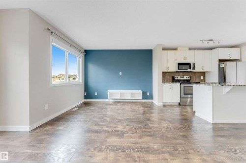 Open-concept living area featuring wood-finish flooring, a large window, a kitchen with white cabinetry, stainless steel appliances, and a breakfast bar with a light-colored countertop - 3340 15A Avenue Nw, Edmonton, AB 