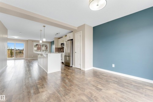 Open-concept layout featuring wood-finish flooring, a kitchen island with pendant lighting, stainless steel appliances, and a private balcony - 3340 15A Avenue Nw, Edmonton, AB 