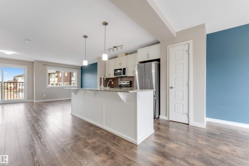 Open-concept living space featuring wood-finish flooring, a kitchen island with a granite-style countertop, stainless steel appliances, and a sliding glass door to a balcony - 3340 15A Avenue Nw, Edmonton, AB 