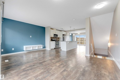 Open-concept living space featuring wood-finish flooring, a built-in media console, and a kitchen with white cabinetry, stainless steel appliances, and a central island with pendant lighting - 3340 15A Avenue Nw, Edmonton, AB 