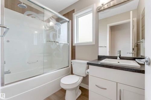 Bathroom featuring a shower-tub combination with sliding glass doors, a window, a single vanity with a dark countertop, and a built-in mirror with integrated lighting - 3340 15A Avenue Nw, Edmonton, AB 