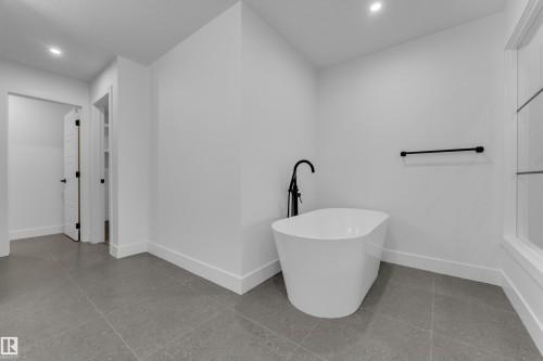 Freestanding soaking tub with a matte black floor-mounted faucet, gray large-format tile flooring, white walls, and a recessed ceiling light - 236 Linden Loop, Leduc, AB 