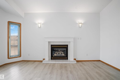 Gas fireplace with a white mantel, two wall-mounted light fixtures, wood-finish flooring, and a tall window with wood trim - 2915 151A Avenue Nw, Edmonton, AB 