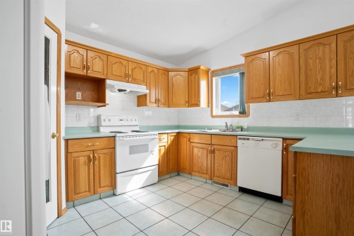 Kitchen featuring wood cabinetry, white appliances, white tile backsplash, a window, and tile flooring - 2915 151A Avenue Nw, Edmonton, AB 