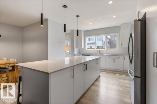 Modern kitchen featuring a spacious center island with a light-toned countertop, white cabinetry with contemporary hardware, stainless steel refrigerator, recessed lighting, and wood-finish flooring - 7113 177A Avenue Nw, Edmonton, AB - Indoor Photo Showing Kitchen With Upgraded Kitchen