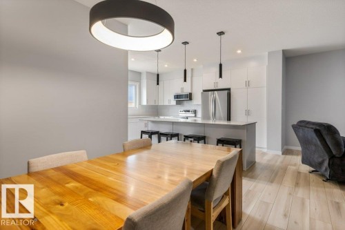 Open-concept living space featuring light wood-finish flooring, a contemporary dining area with a circular pendant light, and a modern kitchen with white cabinetry, stainless steel appliances, and a central island with bar seating - 7113 177A Avenue Nw, Edmonton, AB - Indoor Photo Showing Dining Room
