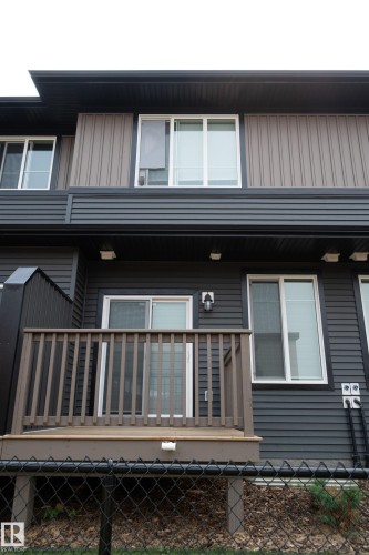 Rear exterior featuring a ground-level deck with wood-finish railing, a sliding glass door, white-framed windows, and dark-toned horizontal siding with vertical panel accents - #64 2710 66 St Sw Sw, Edmonton, AB 