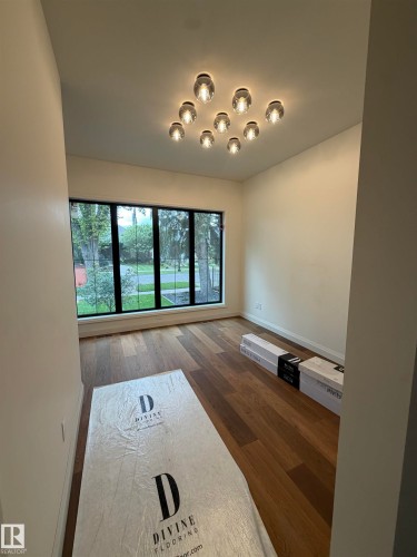 Bright, light-filled room showcasing expansive multi-pane windows framing natural outdoor scenery, warm wood-finish flooring, crisp white walls, and a modern ceiling fixture with integrated pendant lighting - 10223 130 Street Nw, Edmonton, AB 