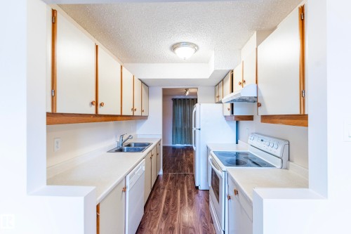 Galley kitchen featuring white cabinetry, light countertops, wood-finish flooring, a double basin sink, and a full suite of white appliances - #4 3221 119 St Nw, Edmonton, AB 