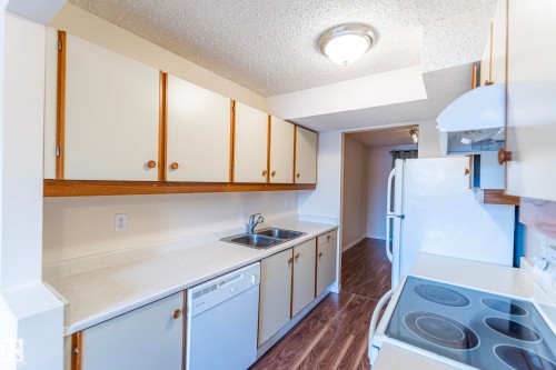 Compact kitchen featuring white cabinetry with wood trim, a stainless steel double sink, white countertops, and wood-finish flooring - #4 3221 119 St Nw, Edmonton, AB 