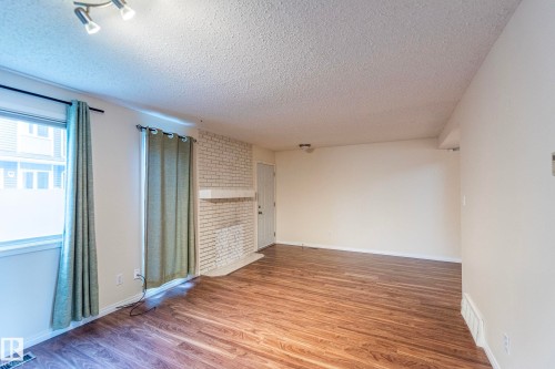 Living area featuring wood-finish flooring, a white brick fireplace, and a large window - #4 3221 119 St Nw, Edmonton, AB 