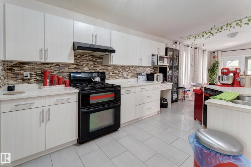 White shaker-style cabinetry with silver-tone hardware, a mosaic tile backsplash, light-toned countertops, a black gas range with a matching range hood, and light-toned tile flooring - 3519 22 Street Nw, Edmonton, AB 