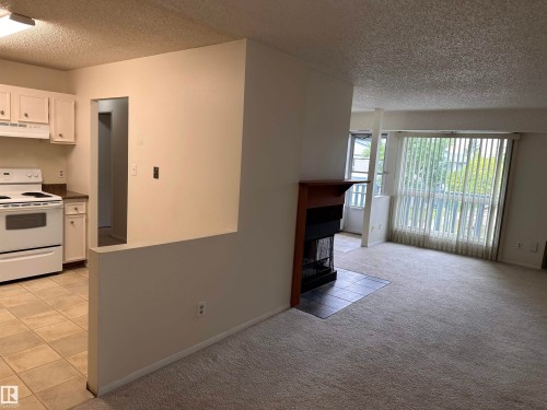 Living area with a fireplace featuring a wood mantle and tile hearth, complemented by a large window with vertical blinds, and soft carpet flooring - #132 2703 79 St Nw, Edmonton, AB 