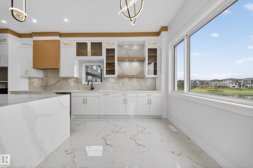 Gourmet kitchen featuring a waterfall island, marble-style flooring, white cabinetry, a wood-finish range hood, and a large window - 8737 Mayday Lane Sw, Edmonton, AB 