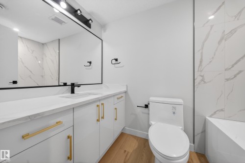 Wood-finish flooring, white vanity cabinetry with gold-tone hardware, rectangular undermount sink with matte black faucet, full-width framed mirror, and large format wall tiling - 8737 Mayday Lane Sw, Edmonton, AB 