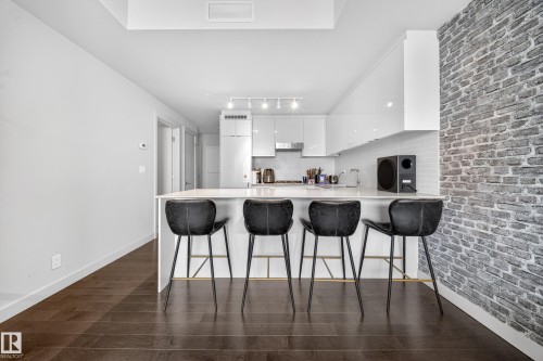 Kitchen island with a white countertop and seating, dark wood-finish flooring, white flat-panel cabinetry, stainless steel range hood, and a textured brick-pattern wall - #4007 10360 102 St Nw, Edmonton, AB 