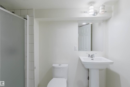Bathroom featuring a walk-in shower with frosted glass enclosure, white subway tile surround, pedestal sink with chrome faucet, wall-mounted mirror, and a two-light vanity fixture - 2439 112 Street Nw, Edmonton, AB - Indoor Photo Showing Bathroom