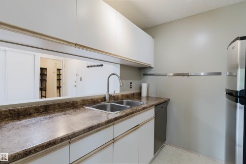 Upper and lower white cabinetry, double basin stainless steel sink with a gooseneck faucet, composite countertop, and stainless steel refrigerator - #316 17109 67 Av Nw, Edmonton, AB 