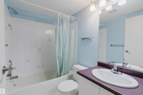 Vanity with integrated sink and chrome faucet, large wall mirror, built-in bathtub with shower, white tile surround with decorative inlays, and overhead lighting fixture - #316 17109 67 Av Nw, Edmonton, AB 