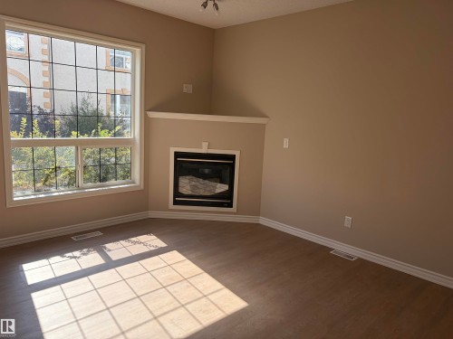 Wall-mounted fireplace with mantel, expansive window with grid, wood-finish flooring, and crown molding - #100 13825 155 Av Nw, Edmonton, AB 