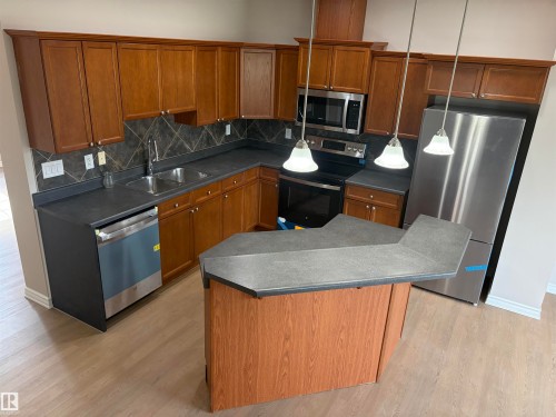 Kitchen featuring wood-finish cabinetry, stainless steel appliances, a center island with a breakfast bar, dark countertops, and wood-finish flooring - #100 13825 155 Av Nw, Edmonton, AB 