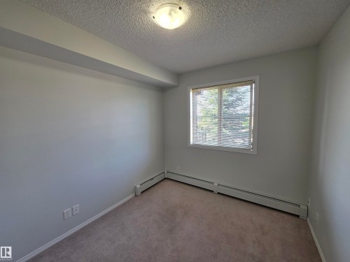 Bright secondary bedroom with carpet flooring, large window with natural light, and baseboard heating - #206 12045 22 Av Sw, Edmonton, AB 