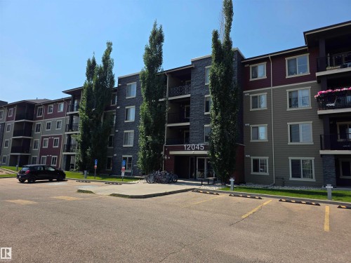 Multi-story building featuring stone-clad and panel siding accents, multiple private balconies, and a paved parking area - #206 12045 22 Av Sw, Edmonton, AB 
