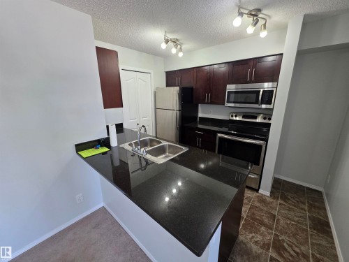 Kitchen featuring dark wood-finish cabinetry, black countertops, stainless steel appliances, and a double basin sink - #206 12045 22 Av Sw, Edmonton, AB 