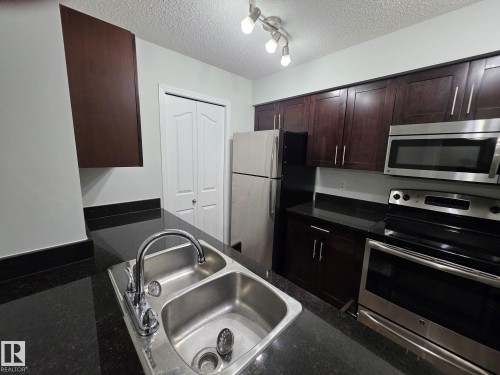 Kitchen featuring dark wood-finish cabinetry, black countertops, stainless steel appliances, and a double basin sink with chrome faucet - #206 12045 22 Av Sw, Edmonton, AB 