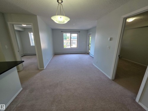 Carpeted living space with a window and exterior door, illuminated by an overhead semi-flush mount light fixture - #206 12045 22 Av Sw, Edmonton, AB 