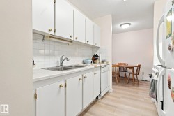 White cabinetry with gold-tone hardware, a double basin stainless steel sink with a chrome faucet, a white tile backsplash, and light wood-finish flooring - 