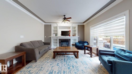 Living area featuring a stone-surround fireplace with built-in shelving, wood-finish flooring, a ceiling fan, and large windows - 1232 Cunningham Drive Sw, Edmonton, AB 