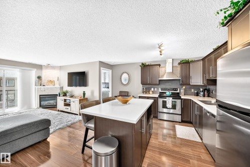 Open-concept living area with wood-finish flooring, a kitchen island with seating, stainless steel appliances, and a fireplace - #418 279 Suder Greens Dr Nw, Edmonton, AB 