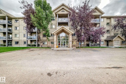 Multi-story condominium building featuring a stone and stucco exterior, prominent entryway, multiple balconies with glass railings, and surrounding paved parking - #418 279 Suder Greens Dr Nw, Edmonton, AB 