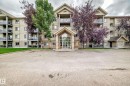 Multi-story condominium building featuring a stone and stucco exterior, prominent entryway, multiple balconies with glass railings, and surrounding paved parking - #418 279 Suder Greens Dr Nw, Edmonton, AB 