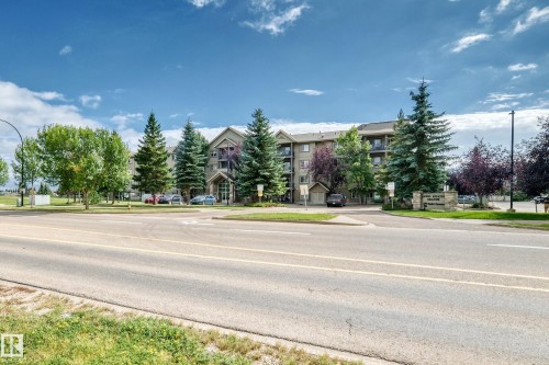 Multi-story building featuring balconies, mature trees, and an asphalt parking area - #418 279 Suder Greens Dr Nw, Edmonton, AB 