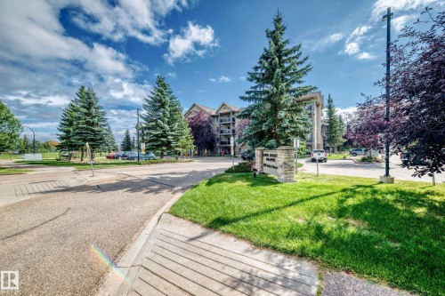 Stone-clad entrance sign, mature evergreen trees, and paved parking area - #418 279 Suder Greens Dr Nw, Edmonton, AB 