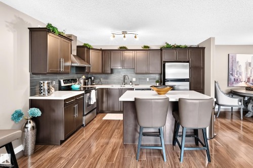 Kitchen featuring wood-finish flooring, dark wood cabinetry, stainless steel appliances, a white countertop island, and a gray subway tile backsplash - #418 279 Suder Greens Dr Nw, Edmonton, AB 
