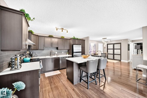 Open-concept kitchen featuring wood-finish flooring, dark wood-finish cabinetry, stainless steel appliances, a white island countertop, and a tile backsplash - #418 279 Suder Greens Dr Nw, Edmonton, AB 