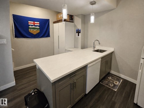 Modern kitchen featuring a spacious white quartz countertop with waterfall edge, white cabinetry with ample storage, stainless steel sink with contemporary faucet, and dark wood-finish flooring throughout - 13309 128 Street Nw, Edmonton, AB 