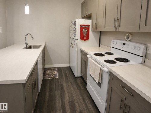 Galley kitchen featuring wood-finish flooring, light-toned countertops, gray cabinetry, a stainless steel sink, and a white electric range - 13309 128 Street Nw, Edmonton, AB 